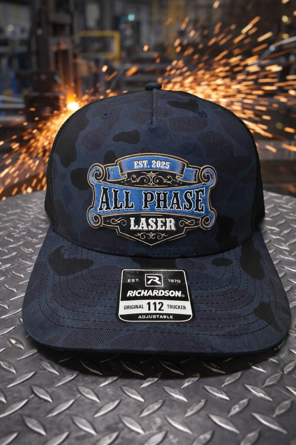 All Phase Laser branded camo trucker hat on diamond plate with welding sparks behind it